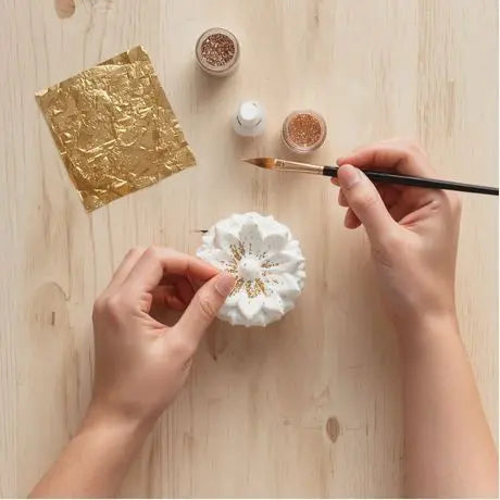 Hände verzieren weißes Blütenobjekt mit Goldglitzer und Pinsel, Goldfolie im Hintergrund