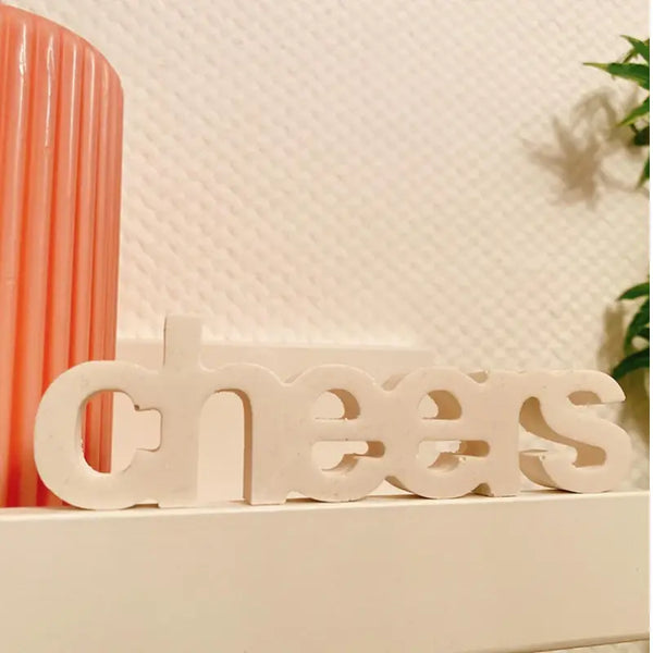 Weißer Cheers-Schriftzug auf Regal mit Rillenkerze und grüner Pflanze